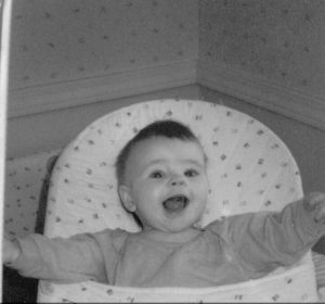 black and white image of a baby in a bouncer, arms outstretched with a smiling face